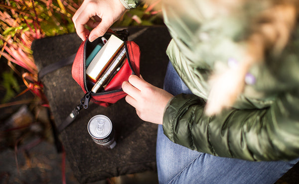 Person wearing a Everyday Cubelet in Mars Red at a Japanese garden. The bag has been taken off and placed next to the person, who has zipped it open to reveal a book, notebook, and pen.